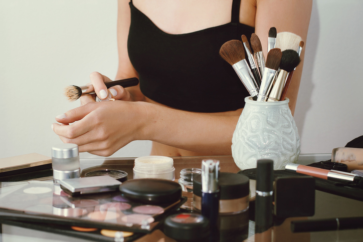 Girl doing make up, pushes concealer on hand. Blurred cosmetics foreground. White background.