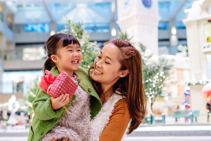 Pretty young mom carrying lovely little daughter who's holding a beautifully wrapped present in her hand, they both smiling joyfully while talking to each other in front of the lighting decoration in the shopping mall